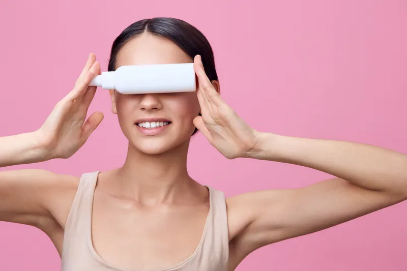 Cheerful young woman holding a white bottle in front of her face against a pink background, promoting hair care and beauty product concepts