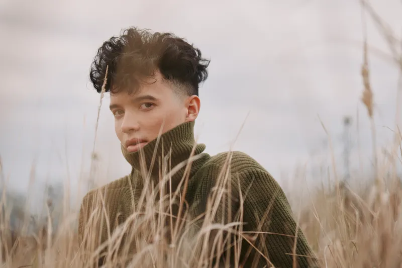 Natural close-up portrait of a young person in tall dry grass, wearing a turtleneck sweater, conveying an authentic, grounded presence and credible, introspective gaze in muted tones and subtle,
