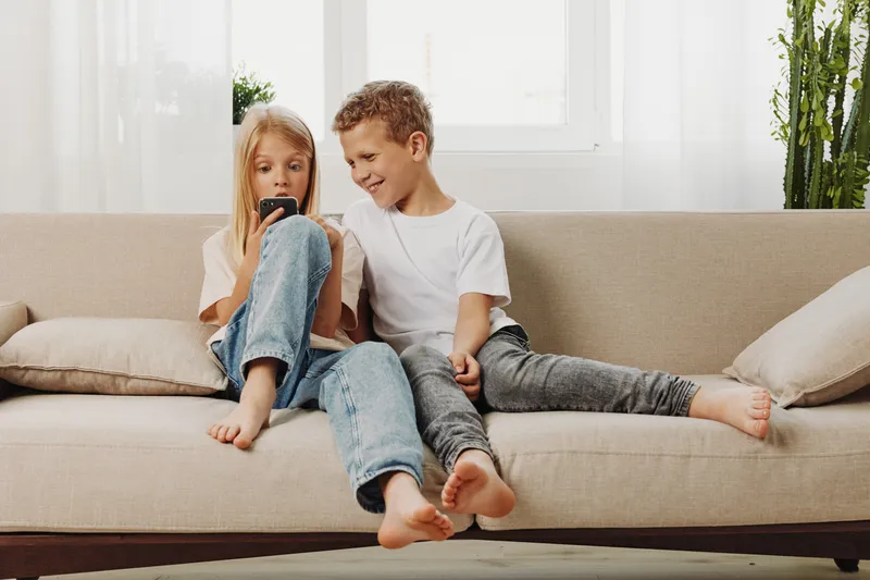 Two children sitting barefoot on beige sofa, boy and girl looking at smartphone together in bright living room with natural light and plants. Concept of childhood, technology, and family bonding.