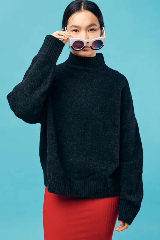 Young woman with long black hair wearing dark sweater and red skirt holding white sunglasses on blue background. Fashion, casual style, confident pose and modern look in studio.