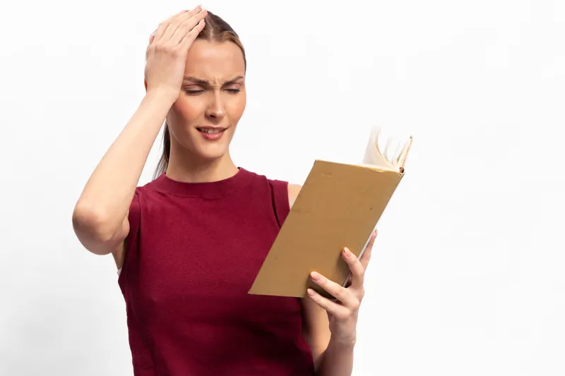 Confused woman in red sleeveless top holds an open book, touching her head with a puzzled expression. Isolated on white background. Concept of frustration, reading difficulty, and concentration.