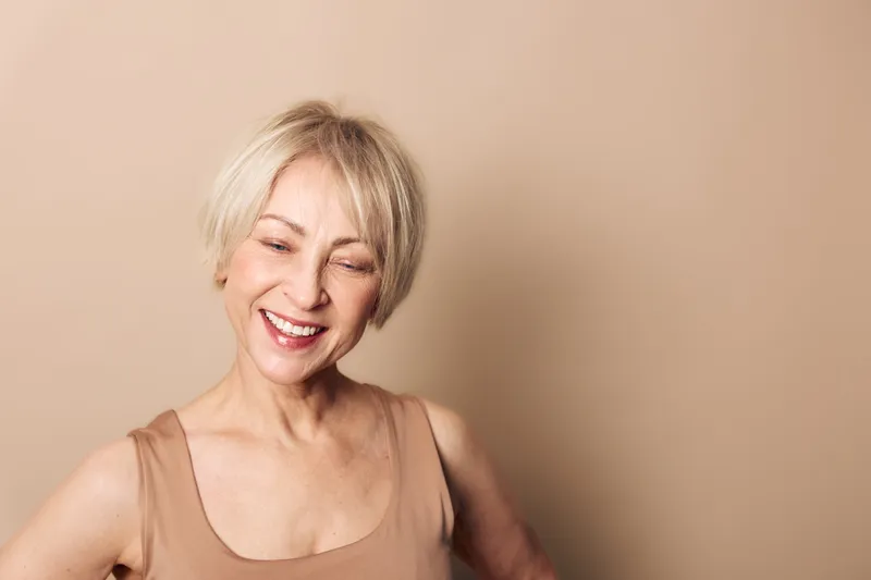 Happy middle aged woman with short blonde hair smiles with closed eyes against beige background. Concept of joy, natural beauty, positivity, and confident mature female portrait.