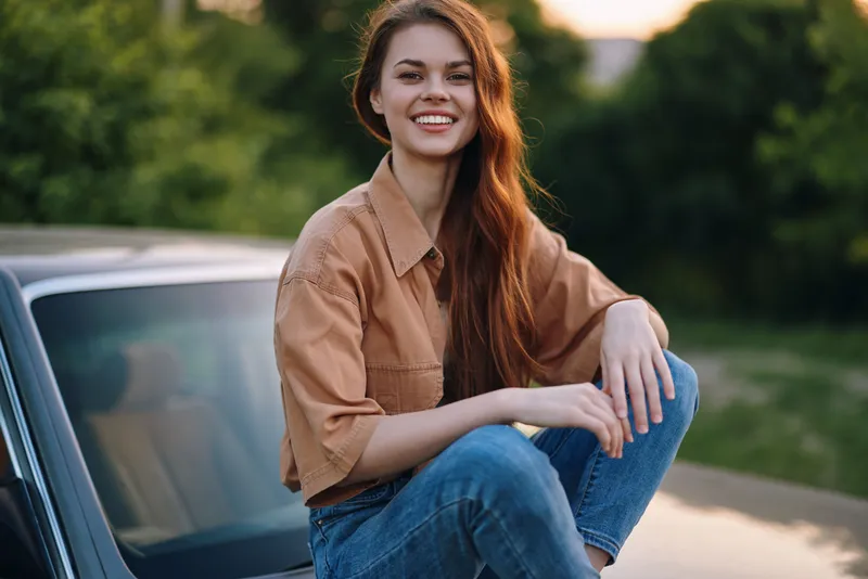 Young woman in casual outfit smiling, sitting on a car, surrounded by greenery, exuding happiness and relaxation, perfect for lifestyle and summer themes.