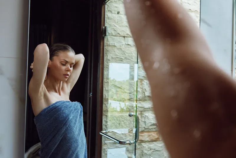 woman in towel adjusting hair near glass shower door with water droplets, indoor bathroom scene, natural light and calm mood, relaxed young adult