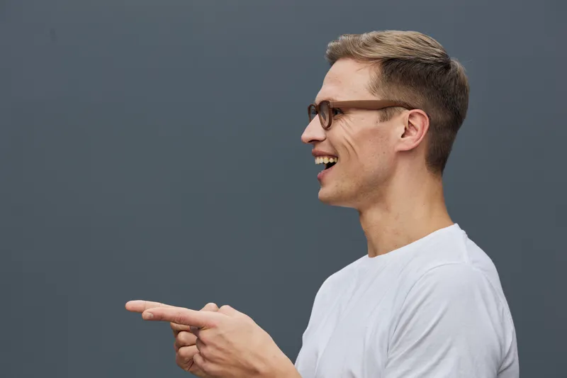 Smiling young man with glasses wearing white t-shirt, pointing sideways with hand, looking to the right, cheerful expression, casual style, isolated on plain dark background, studio portrait. People l