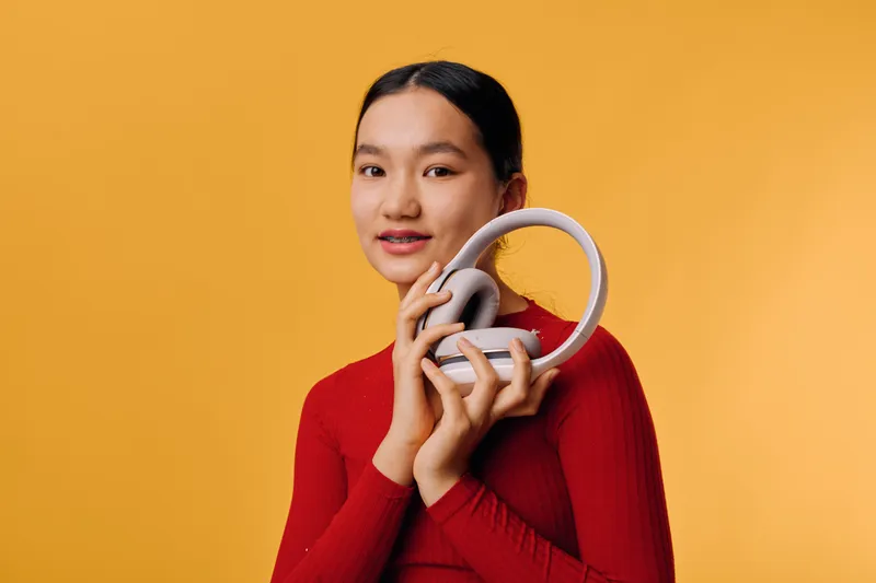 Young woman with headphones standing against yellow background looking at camera holding device in red sweater in studio. Portrait of female person with casual style and modern audio gadget.