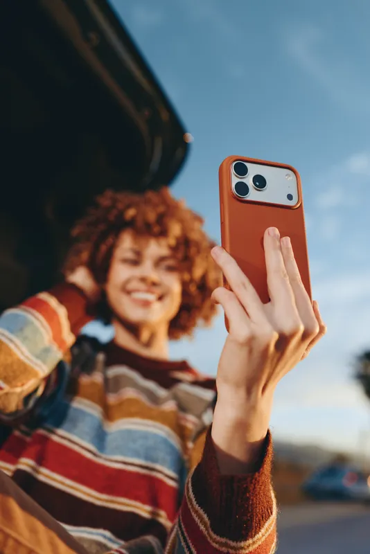 Woman lifestyle outdoors, smartphone selfie, curly hair, smiling hands gesture, blue sky background, warm natural light, soft breeze, casual sweater stripes