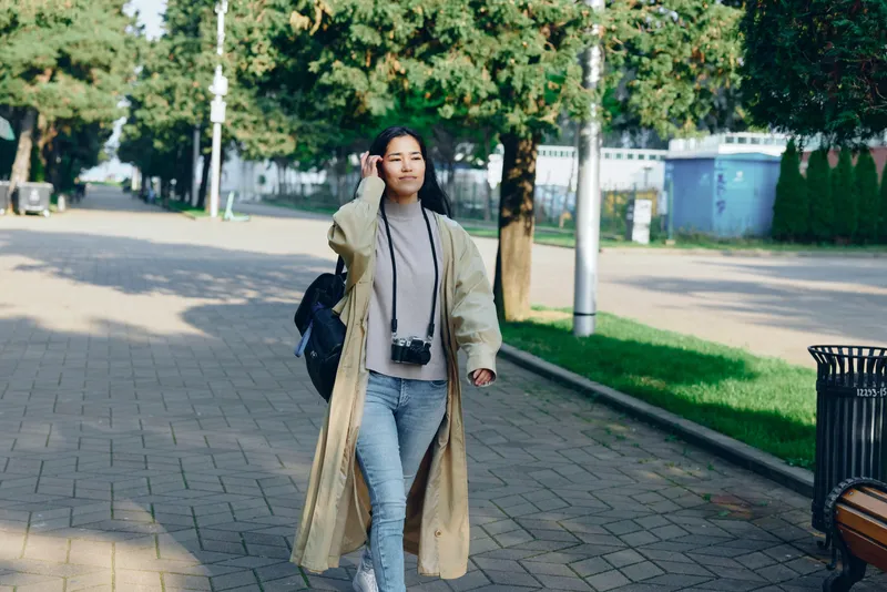 Woman walking outdoors with a camera hanging from her neck, wearing a long coat and jeans, on a tree lined street in daylight, casual urban stroll