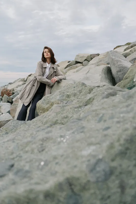 Young woman in light coat and black pants leans on large rocks outdoors under cloudy sky. Concept of fashion, nature, and casual style with a moody atmosphere.