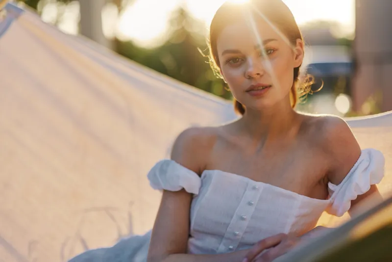 Young woman in a white summer dress relaxing outdoors, showcasing tranquility and joy in a sunlit hammock surrounded by greenery