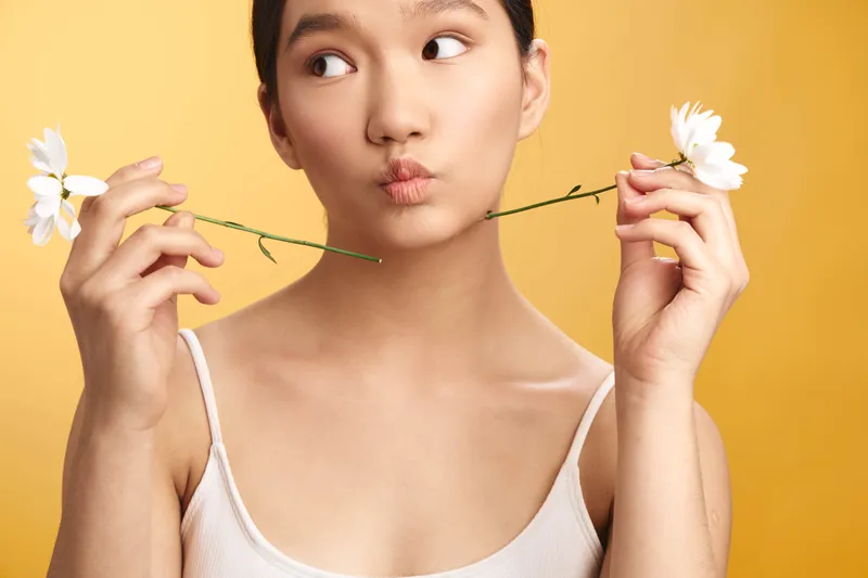 Asian woman holds flowers close to her face against a bright warm backdrop, creating a playful yet elegant mood. She smiles softly, exuding calm and charm while the floral props add a fresh,