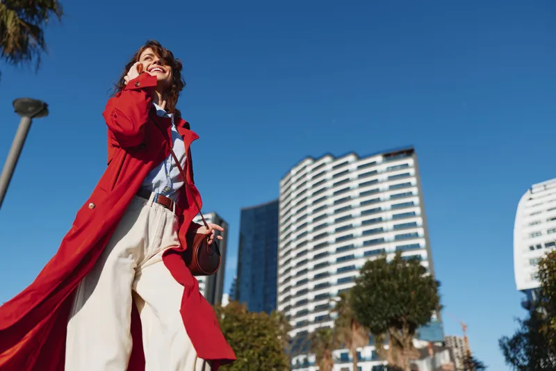 A stylish woman in a red coat strides through a sunny city street, looking up with confidence, capturing a candid moment of urban fashion and energy.