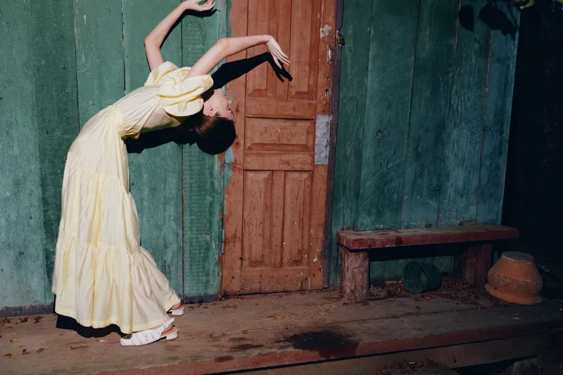 Dancer in a pale yellow dress arches gracefully beside a rustic doorway, performing an expressive movement outdoors against a weathered green wall.