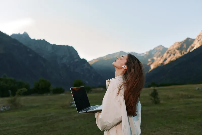 A cheerful professional woman stands outdoors in a scenic valley with mountains, smiling as she holds a laptop, exuding optimism, focus, and productive energy.