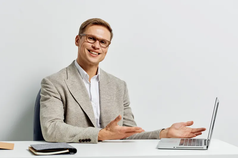 Smiling man in formal attire sitting at desk with open laptop, gesturing with hands during presentation or discussion, professional office background, bright lighting, confident demeanor, business con