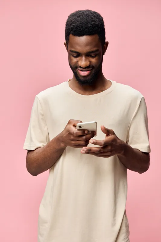 Young African man enjoying tech, smiling while using smartphone on pink background, showcasing modern lifestyle and connectivity