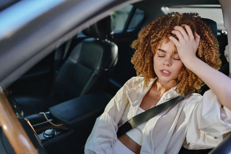tired woman with curly hair sitting in car, wearing white jacket, fastening seatbelt, showing fatigue and stress, natural light inside vehicle interior