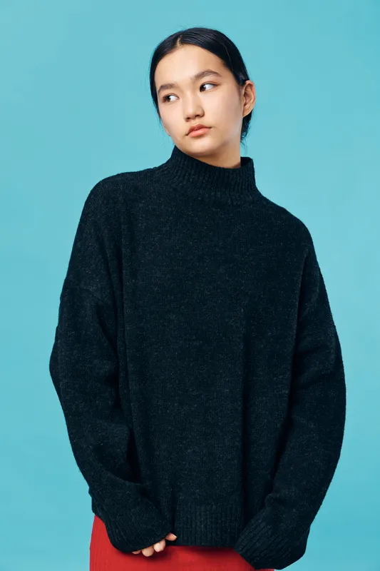 Young woman wearing black sweater standing against blue background looking sideways with thoughtful expression and minimal makeup with natural hairstyle and casual pose.