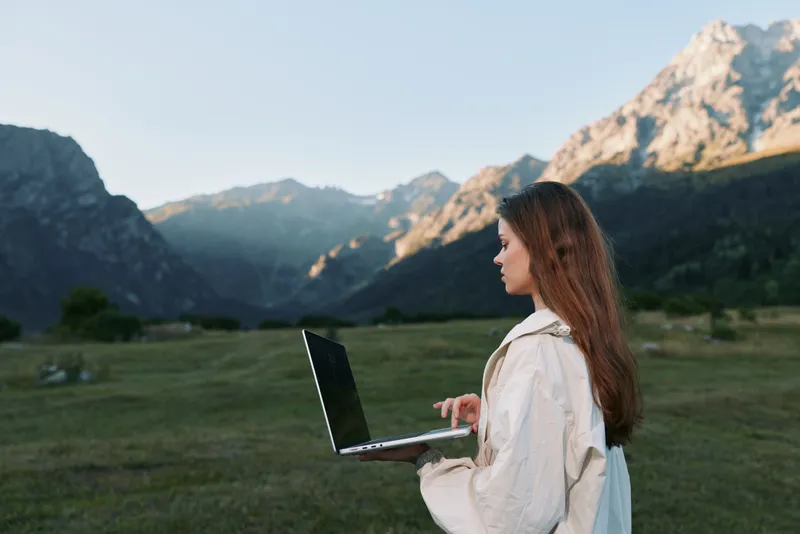 A focused woman works on a laptop in a scenic mountain field, enjoying fresh air and solitude while staying productive in an outdoor remote work setting.