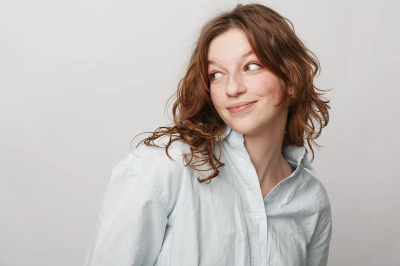 Young woman with curly brown hair wearing a white shirt looks sideways with a slight smile on a light background. Concept of natural beauty, casual style, and positive mood.