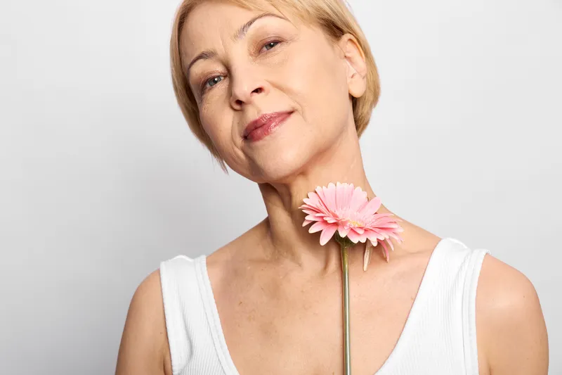 Woman portrait senior blonde with pink flower in studio closeup, beauty calm expression, soft light on skin, sleeveless white top, neutral studio background