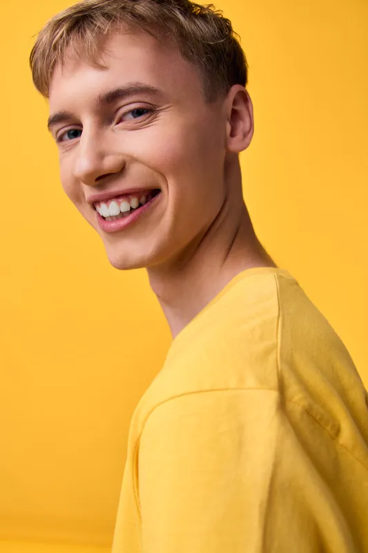 Smiling young man in a yellow shirt stands against a bright background, showcasing casual fashion, happy expression, and a positive mood in a studio setup