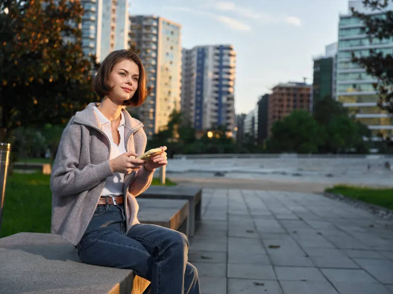 A cheerful professional sits outdoors in a modern city plaza, texting on a smartphone. She smiles warmly, creating a positive, energetic business vibe in a bright urban environment.