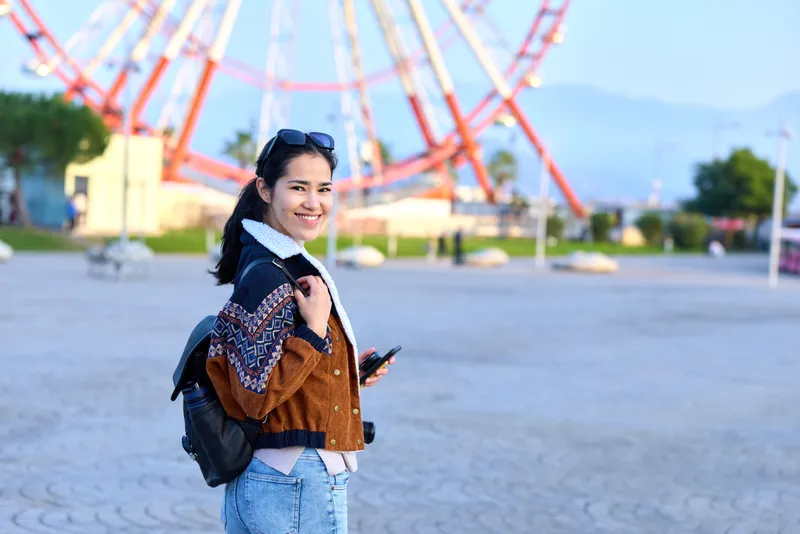 Female traveler with backpack and smartphone in outdoor amusement park, smiling near ferris wheel, casual sweater and jeans, sunny day, candid exploration.