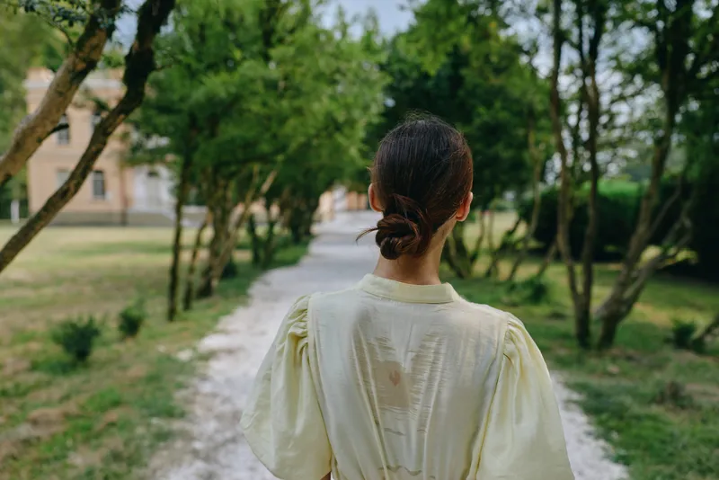 Woman in a pale yellow dress is seen from behind, walking along a gravel path through a lush park, sunlight filtering through trees, creating a serene outdoor scene.