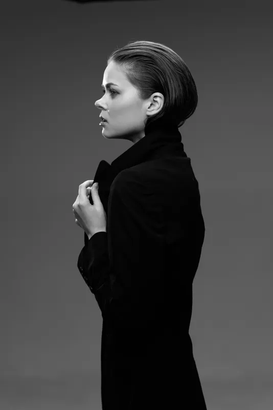 Black and white portrait of a young woman in a dark coat, looking sideways with a serious expression and elegant hairstyle.