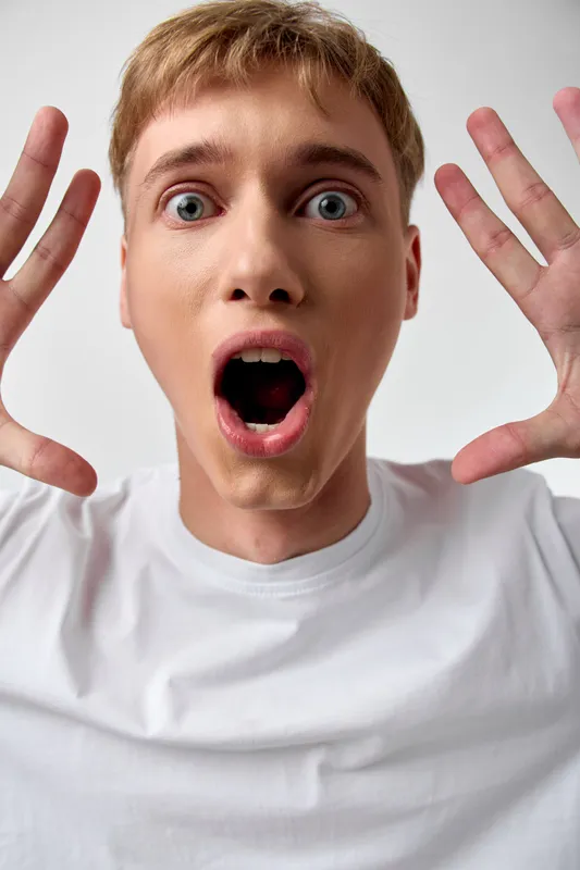 Surprised man with wide eyes and open mouth, hands raised in a dramatic gesture, wearing a white shirt, capturing shock, astonishment and spontaneous emotion in studio setting
