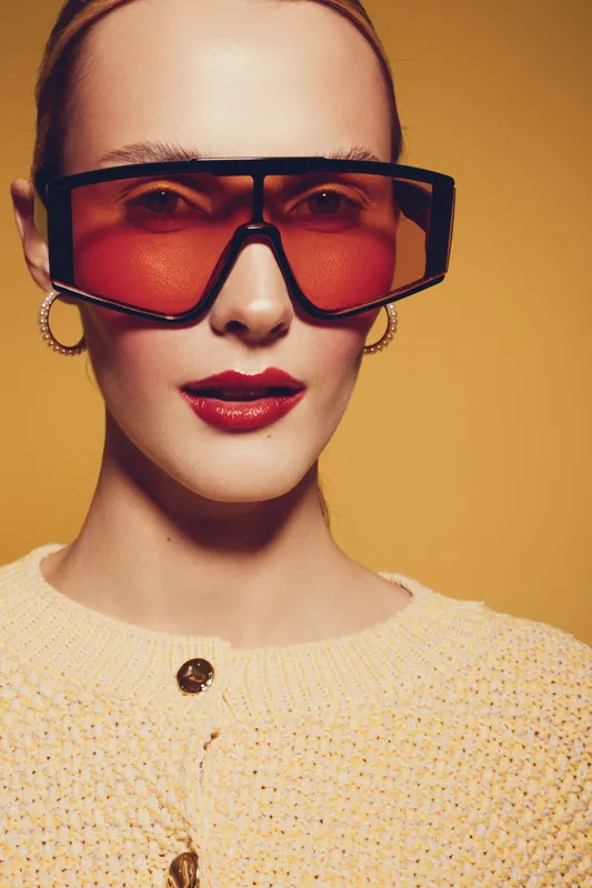 Stylish woman with oversized red sunglasses and hoop earrings poses against yellow background. She wears a textured beige sweater with buttons. Concept of fashion, accessories, and modern style.