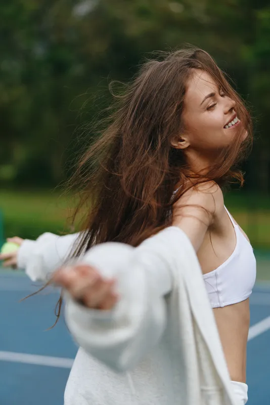 Joyful young woman dancing on a tennis court, wearing sporty attire, embodying freedom and happiness in a vibrant green outdoor setting