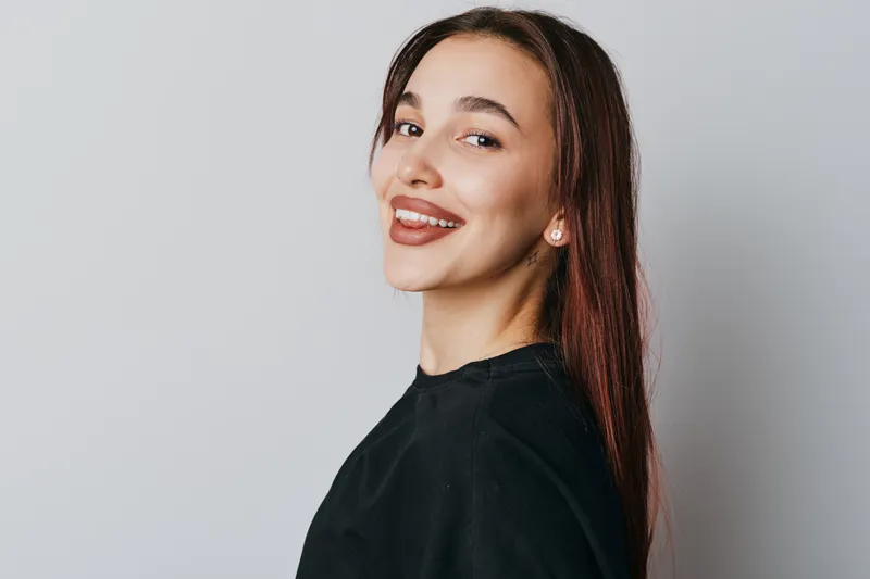 Young woman with long brown hair smiling and posing against white background in black shirt. Portrait of happy female with natural makeup and confident expression looking at camera.