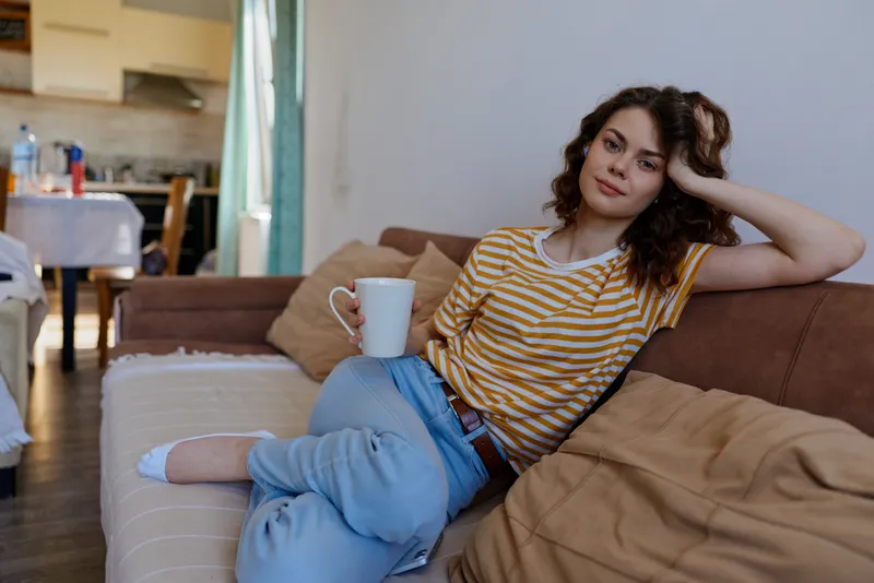 Relaxed woman lounging on a couch at home, striped t shirt, blue jeans, holding a mug. Casual living room scene highlighting comfort, self care, and daily routine. Soft natural lighting creates a