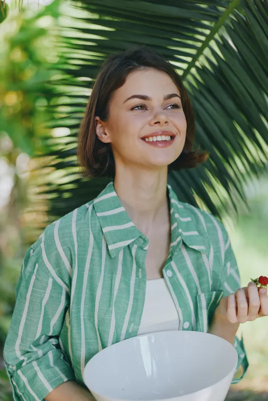 A woman smiles outdoors, wearing a green striped shirt, perched among lush greenery while holding a white bowl, capturing a sunny, relaxed moment of casual fashion and natural charm.