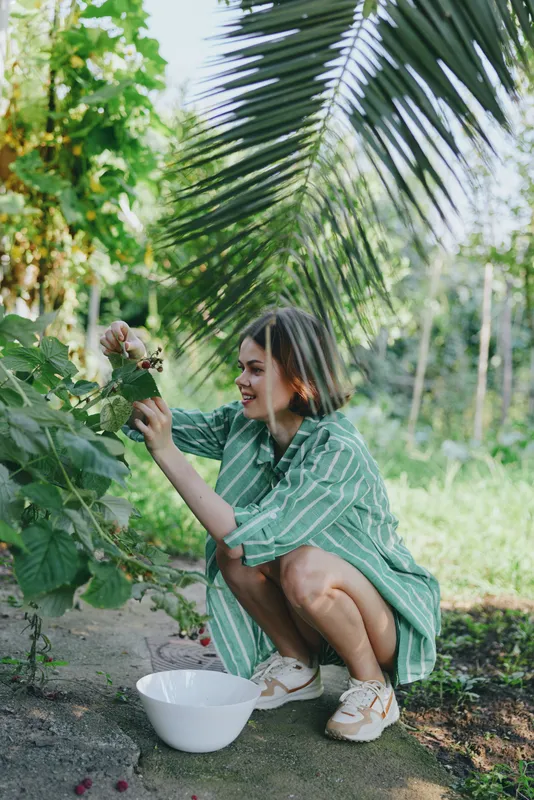 A woman in a green striped dress kneels beside plants, picking flowers into a white bowl, enjoying a sunny garden moment.