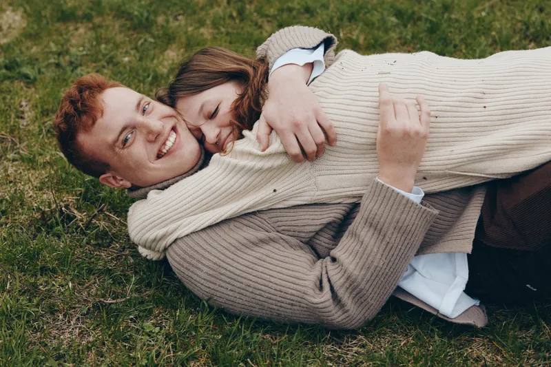 Smiling couple lying on a grassy lawn, embracing each other in a relaxed outdoor scene. Warm natural light highlights affection, happiness, and togetherness in a cozy lifestyle moment.