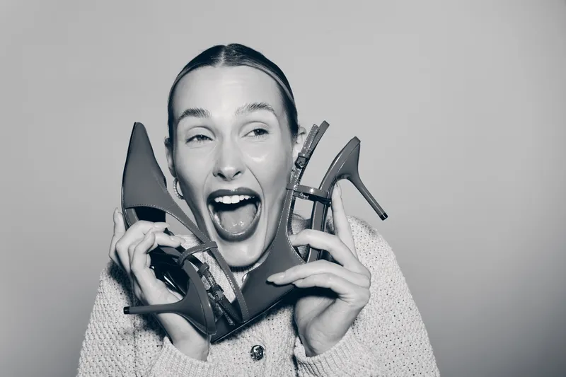 Excited white woman with fair skin holding two high heel shoes near her face, expressing joy and enthusiasm in a casual outfit. Fashion concept.