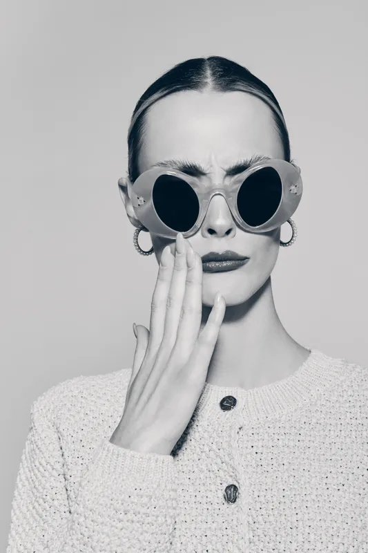Young white woman with pale skin and slicked hair wears stylish round sunglasses and earrings touching face in studio light.