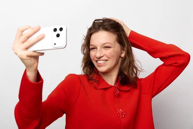 Smiling woman in red sweater takes selfie with smartphone on white background. Casual lifestyle, happiness, and modern technology concept with natural expression and relaxed pose.