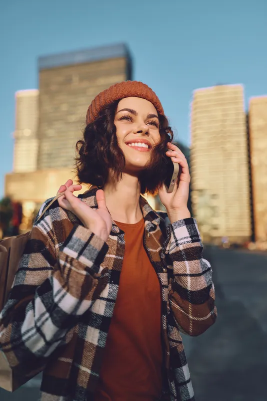 woman smile phone city backpack beanie plaid sunlight candid lifestyle portrait capturing authenticity and emotional storytelling as a young traveler enjoys golden hour glow and mindful living in