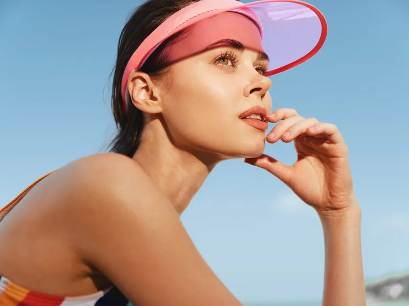 Portrait of a sunlit woman wearing a pink visor, gazing toward the horizon with a relaxed pose, warm glow on skin, and vibrant summer mood.
