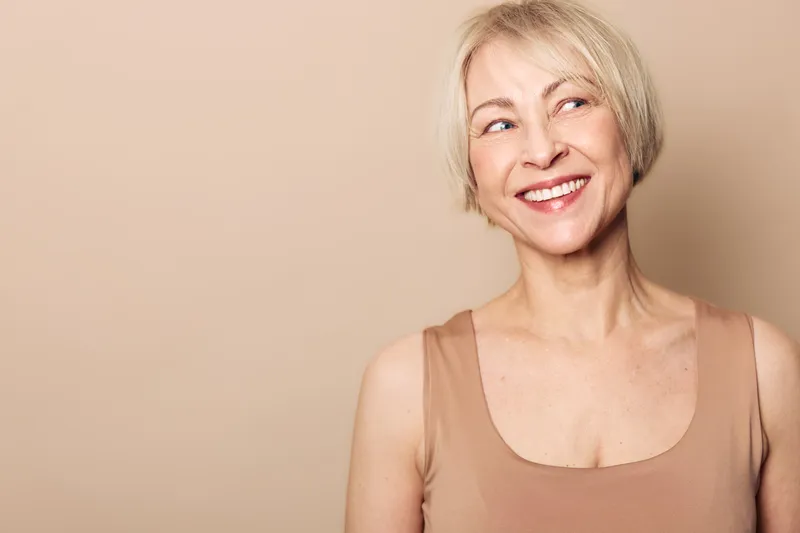 Smiling middle aged woman with short blonde hair wearing beige sleeveless top looks away thoughtfully. Studio portrait with soft warm lighting on neutral background for positive emotions concept.