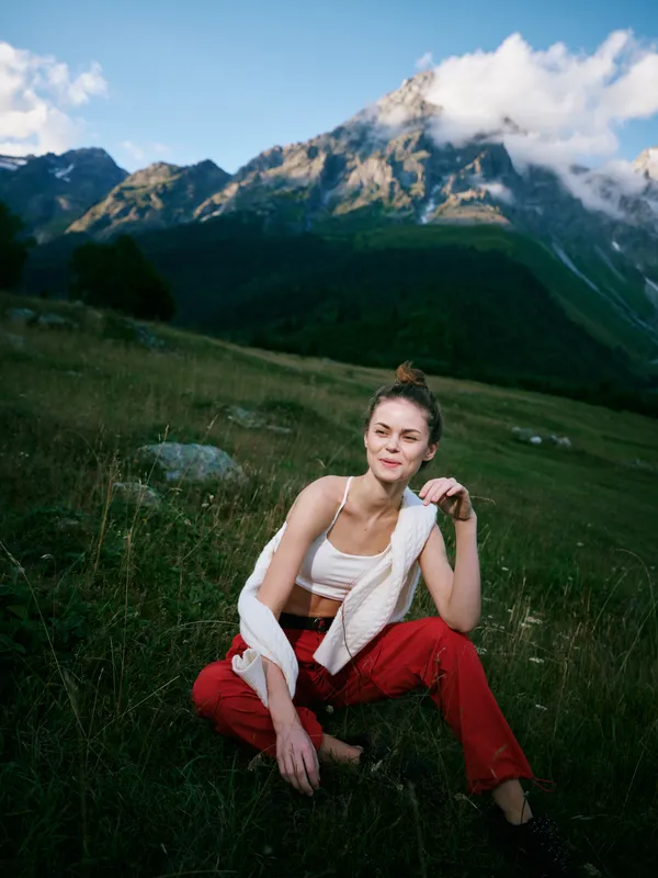 Female sitting in a lush alpine meadow with towering mountains behind, wearing a white top and red pants, a gentle breeze, warm sunlight, and a tranquil outdoor moment in nature