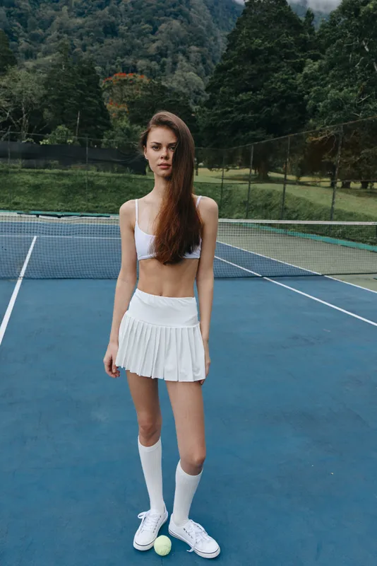Young athletic woman in white sportswear stands confidently on a tennis court with a green backdrop, embodying fitness and determination