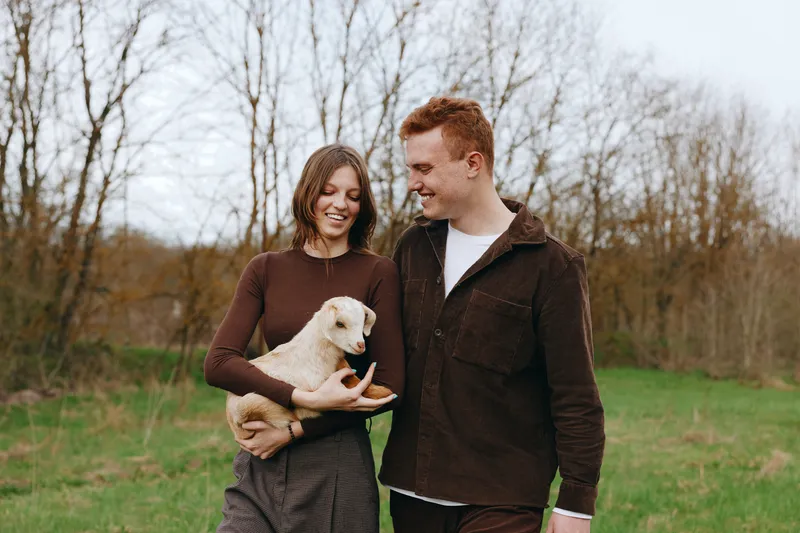 A joyful couple walks outdoors with a playful goat kid in their arms, capturing a warm lifestyle moment in a green field. The scene shows companionship, nature, and gentle animal interaction.