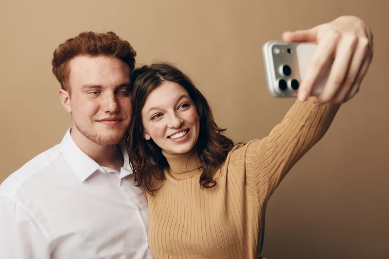 Young couple with red hair and brown hair takes a selfie together on smartphone. They smile and pose in casual clothes against beige background. Concept of friendship and modern technology.