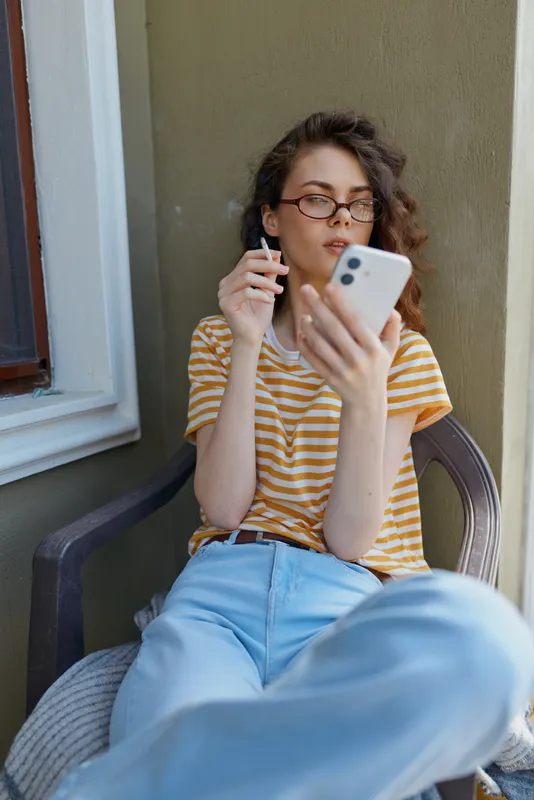 Woman relaxing on a chair at home, looking at smartphone, wearing striped yellow shirt and jeans, casual lifestyle scene with natural light, focused moment showcasing everyday comfort