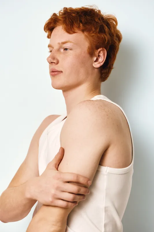 Young man with red hair and freckles in white sleeveless shirt posing sideways against white background with serious expression. Natural light highlights skin texture.